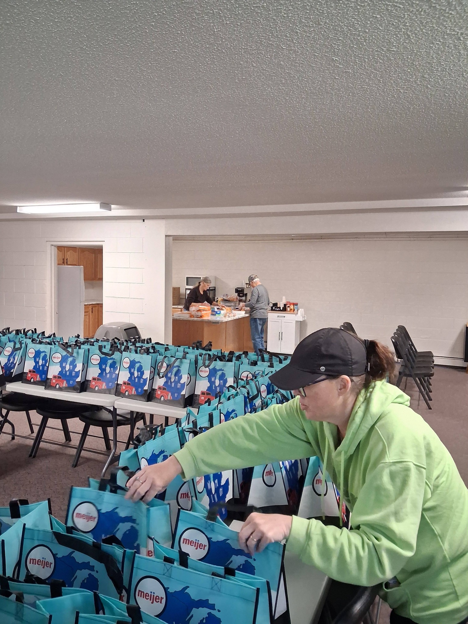 A person in a green jacket and black cap packs turquoise Meijer branded gift bags or promotional items in what appears to a community event or donation distribution space.