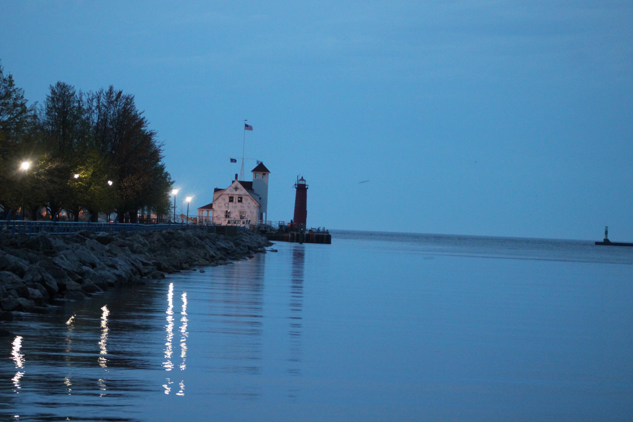 A scenic twilight photograph of a lighthouse and keeper's house on a rocky breakwater, with flags flying and calm blue water reflecting evening lights.
