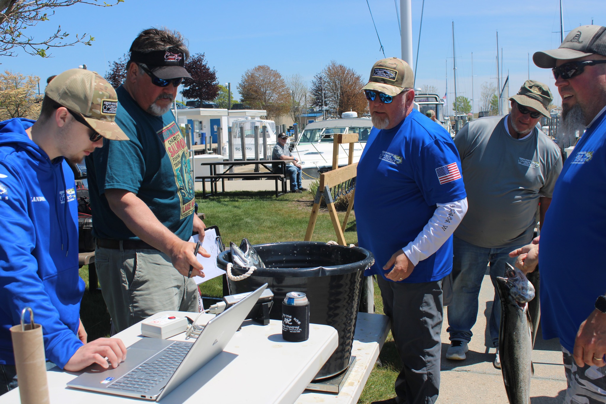 Armed Forces Salmon Assault event with volunteers registering participants at a waterfront venue, featuring a large cooking pot and fresh-caught salmon.