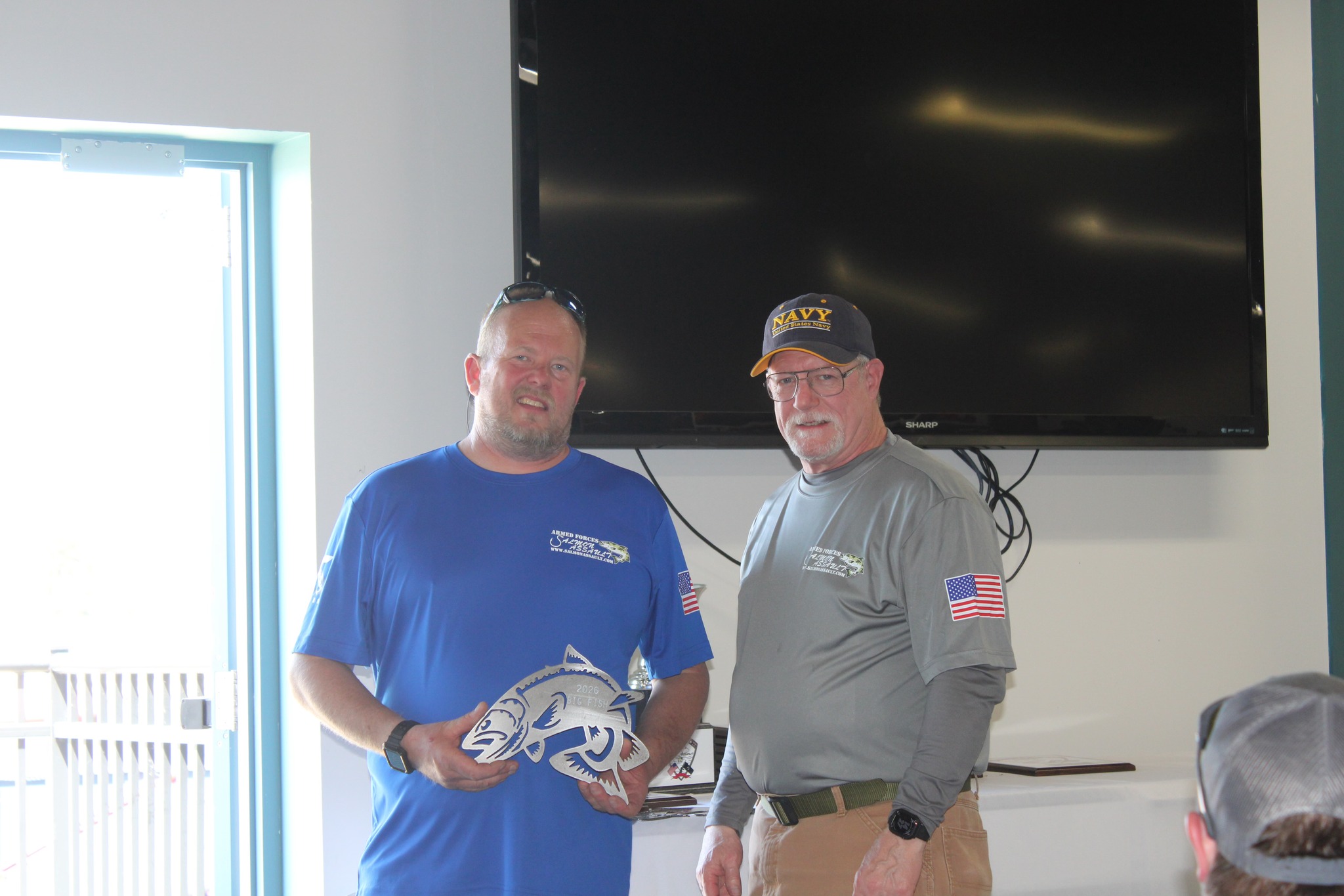 Two men wearing Armed Forces Salmon Assault branded shirts stand together indoors, with one holding a decorative salmon plaque.