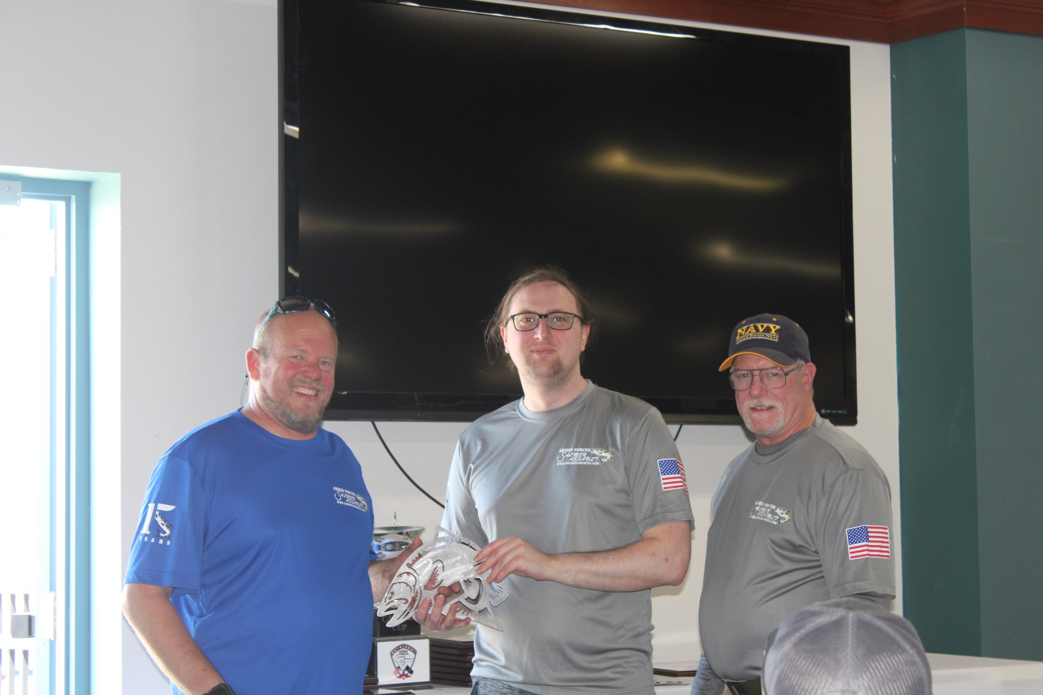 Three men wearing armed-forces-salmon-assault branded shirts pose together indoors, with one holding a decorative fish sculpture.
