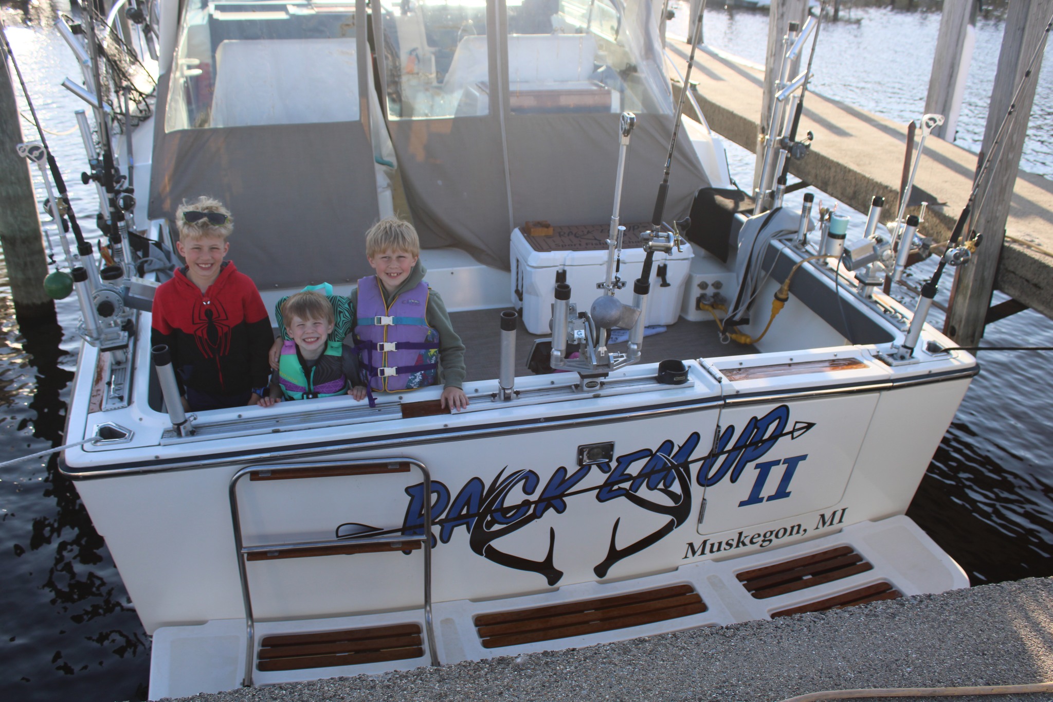 Three children wearing life jackets smile from the deck of the Backerrup II fishing charter boat docked in Muskegon, MI.